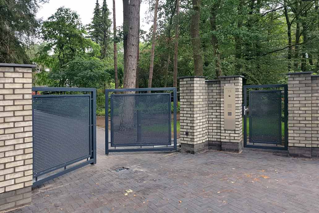 Auf der linken Seite ein Drehflügeltor mit einer Lochblechfüllung zwischen hellen Klinkerpfosten. Rechts eine Personentür aus demselben dunkel beschichtetem Stahl. Im Hintergrund ein parkähnliches Grundstück mit hohen Bäumen.