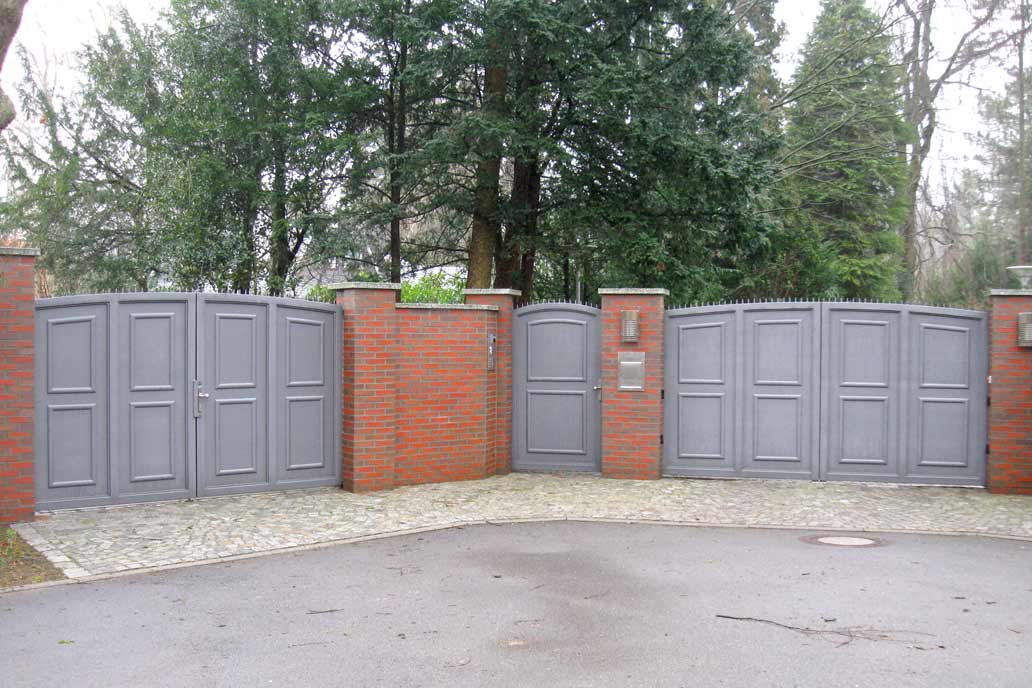 Links und rechts im Bild jeweils ein graues blickdichtes Drehflügeltor zwischen roten Mauerpfosten. In der Mitte eine Personentür aus demselben Material. Im Hintergrund hohe grüne Nadelbäume.